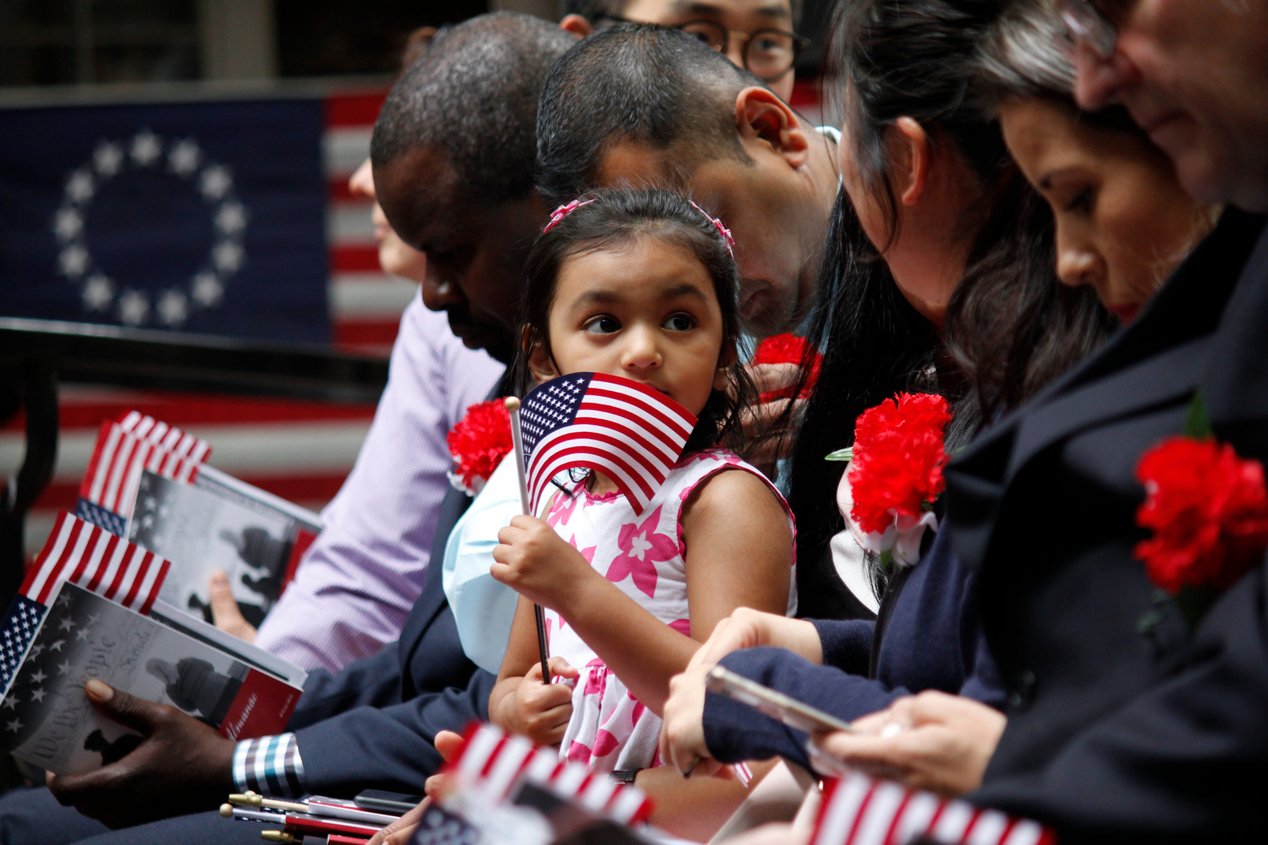 A young girl holds an American flag while sitting among a group of people, including immigrants, at a patriotic event. The girl looks to the side, surrounded by individuals holding flags and wearing red flowers, with a U.S. flag displayed in the background.