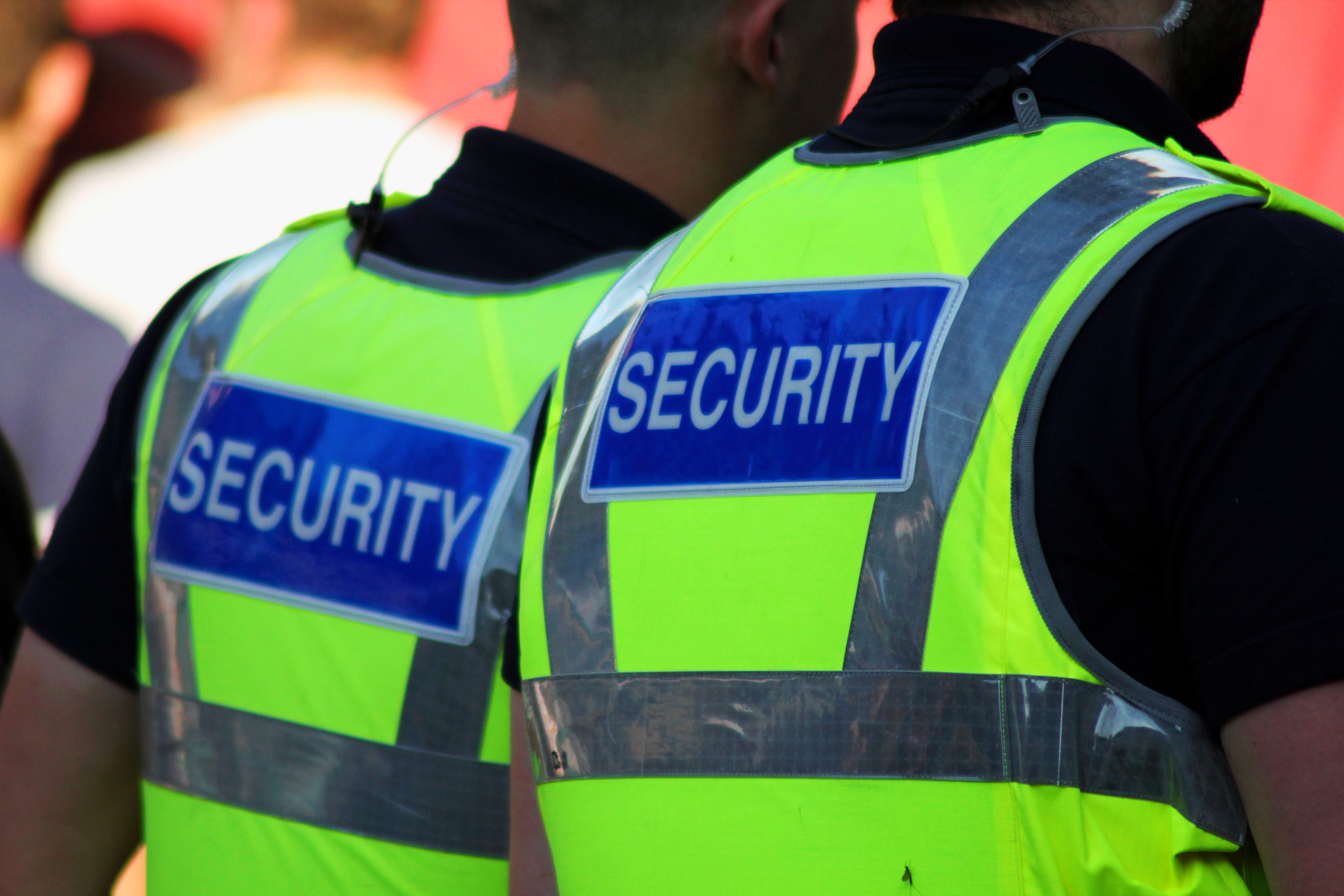 Two male security guards wearing bright yellow reflective vests with the word 'SECURITY' on their backs, standing together during an event or duty.
