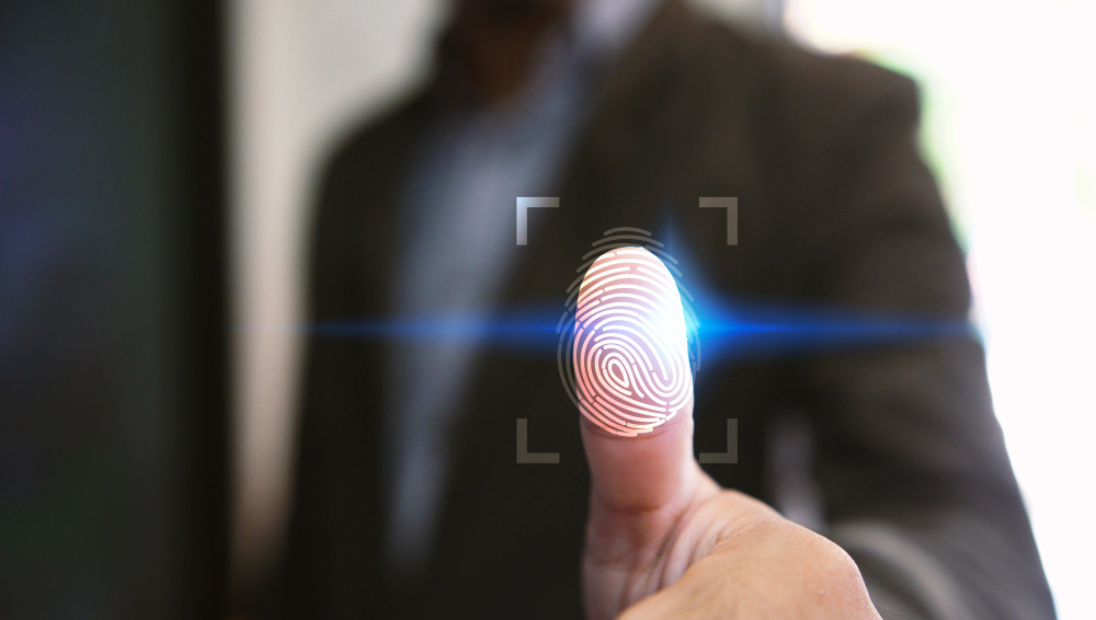 A person wearing a suit is using their thumb to activate a biometric fingerprint scanner. A glowing digital overlay highlights the fingerprint, with a blue light scanning horizontally across it.